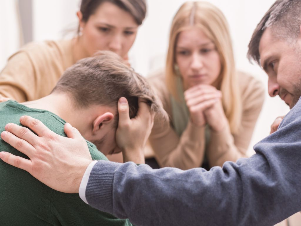 Close-up of a devastated young man holding his head in his hands and friends supporting him
